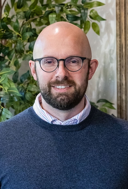 Andrew-Castleberry-Bio-2026 man with glasses, a beard, and a mustache is smiling, wearing a blue sweater over a collared shirt, standing in front of leafy green plants and a framed picture.