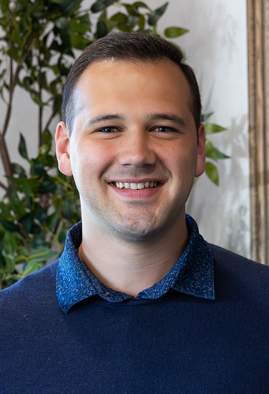 Austin-Rowland-Bio-2026 A young man with short dark hair, wearing a blue patterned-collar shirt and navy sweater, smiles warmly at the camera. Green leafy plants and a frame are visible in the softly lit background.