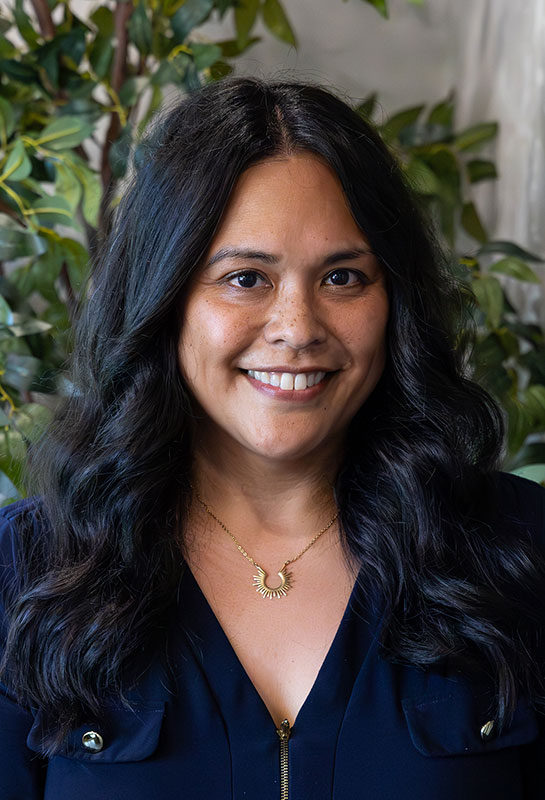 Carol-Taylor-Bio-2026 A woman with long, wavy black hair smiles at the camera. She is wearing a navy blue top and a gold necklace, standing in front of green, leafy plants.