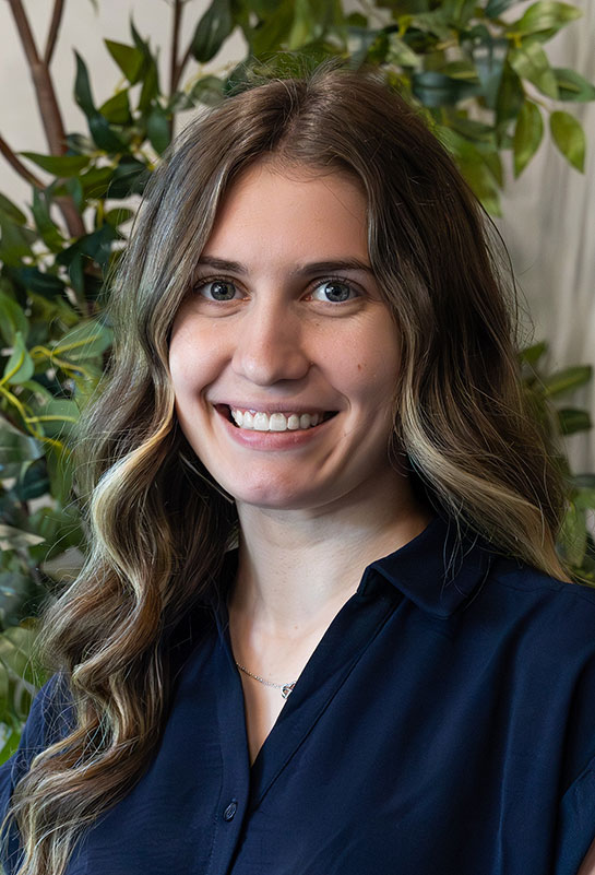 Cloe-Trainer-Bio-2026 A young woman with wavy light brown hair, wearing a navy blue blouse, smiles at the camera. She is standing in front of a leafy, green indoor plant background.