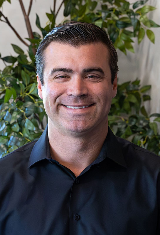 Ken-Bellavance-Bio-2026 A man with short dark hair, wearing a black collared shirt, smiles at the camera. He is standing in front of a leafy green plant background.