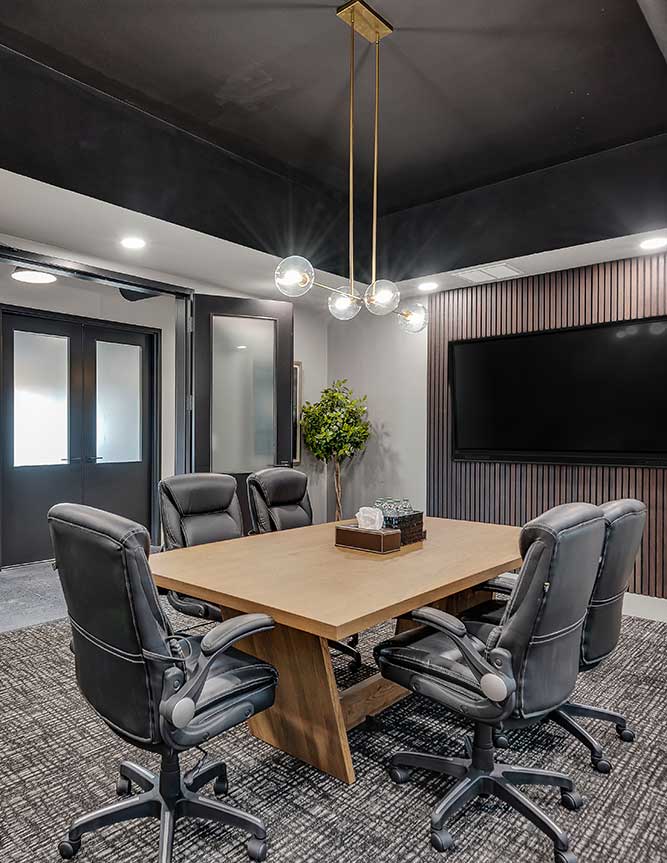 Modern conference room at TruFP financial planning in Cumming, Atlanta, GA, featuring a wooden table, five black leather chairs, a large wall-mounted TV, a decorative plant, and a contemporary light fixture hanging from a dark ceiling.