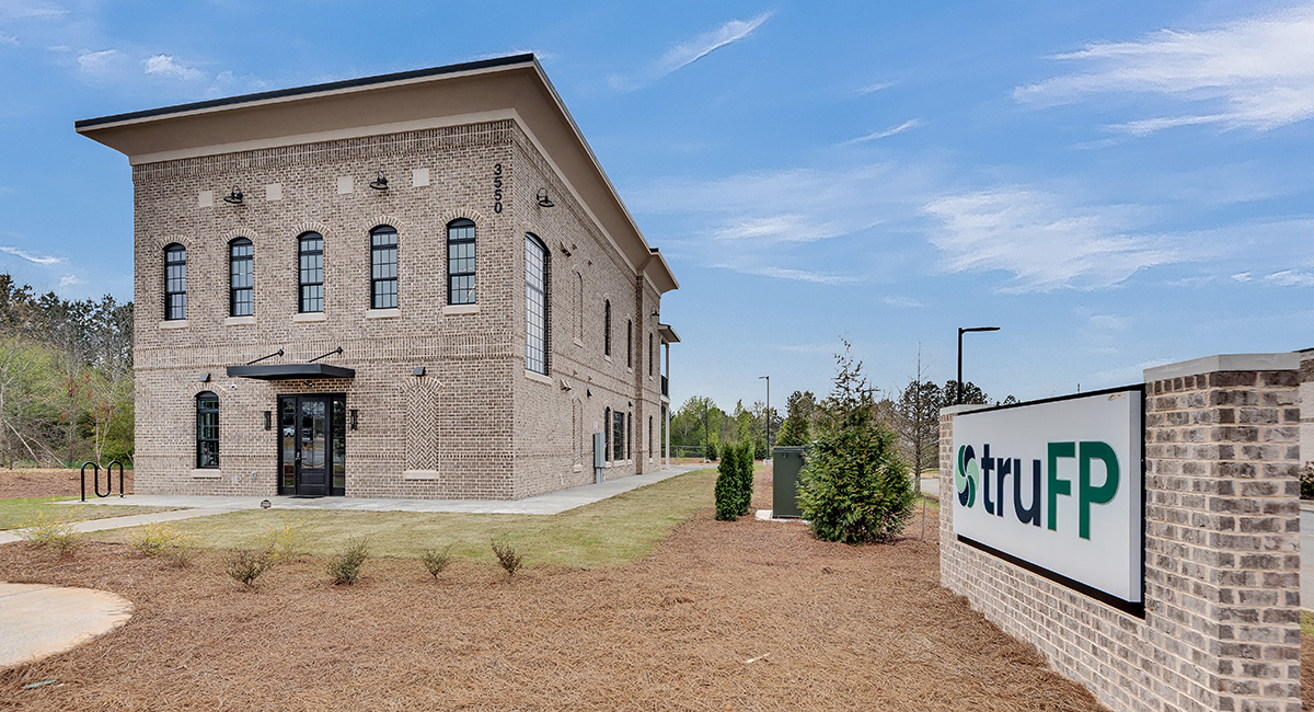 A modern two-story brick building with large windows sits beside a lawn, with a sign in front reading "struFP." The sky is blue and clear, and the area is landscaped with small bushes and pine straw.