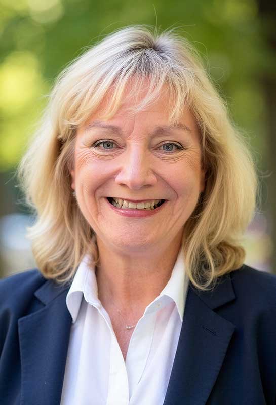 Wendy-Paxton-2026 A smiling woman with blonde hair wearing a dark blazer and white shirt, standing outdoors with a blurred green background.